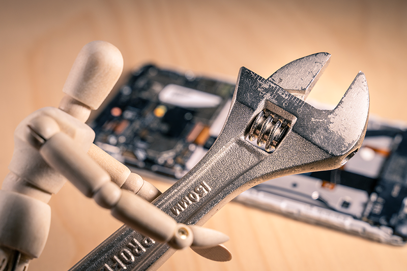 A wooden dummy holding a full-sized wrench in front of a taken-apart phone device