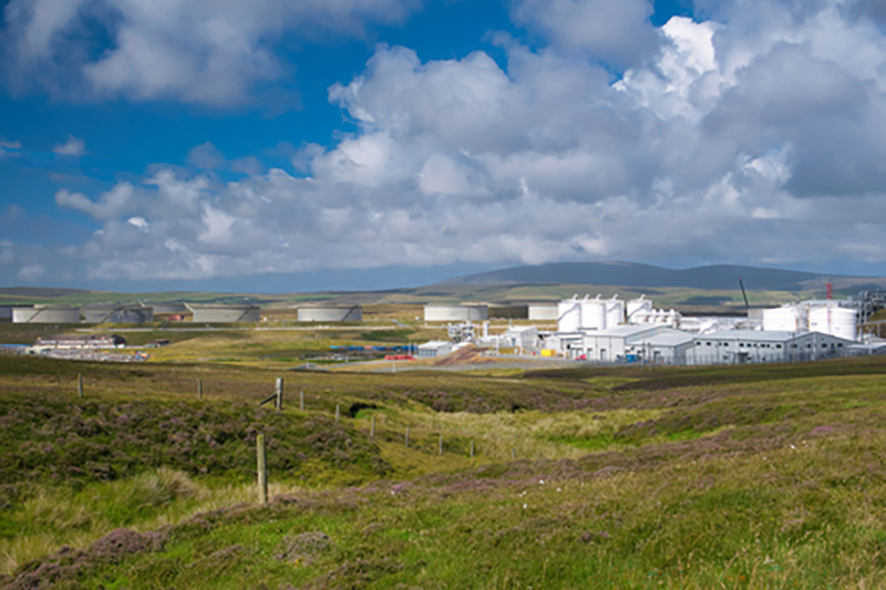 A series of low-lying industrial buildings, including large gas tanks, in the distance of a rolling, treeless landscape