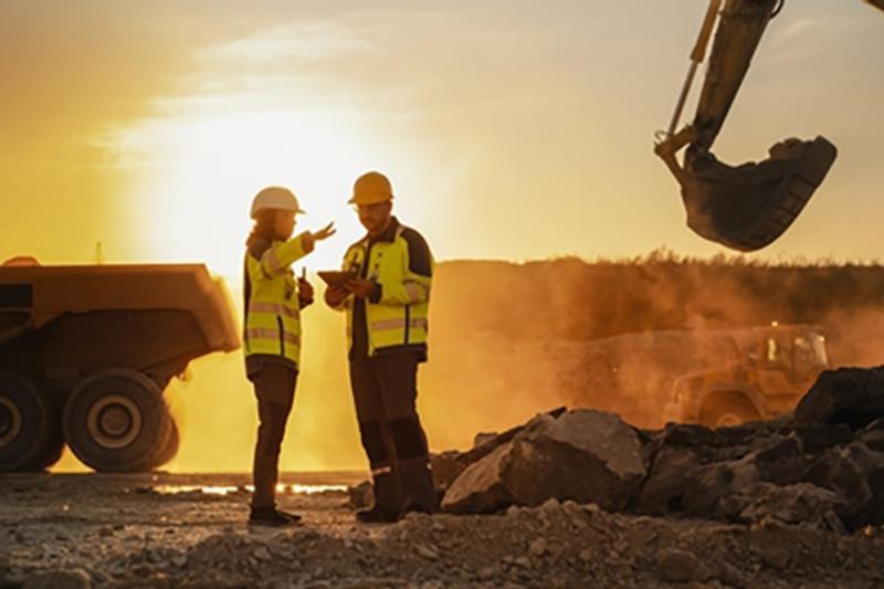 A man and a women on a construction site in the evening sunshine, in hi-vis jackets and helmets, with a truck to one side and a digger the other and stones on the ground