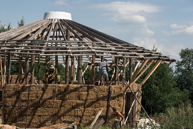 A large wooden-framed circular building being built with straw bale walls by construction workers standing on the top of them