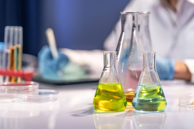 Close-up of colourful chemical liquids in Erlenmeyer flasks on a laboratory table, with a scientist working in the background.