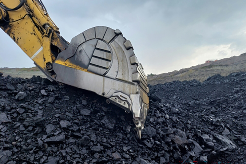 A close up of part of the arm and bucket of an excavator resting on coal in the open air, with grey clouds behind and a slope of a hill