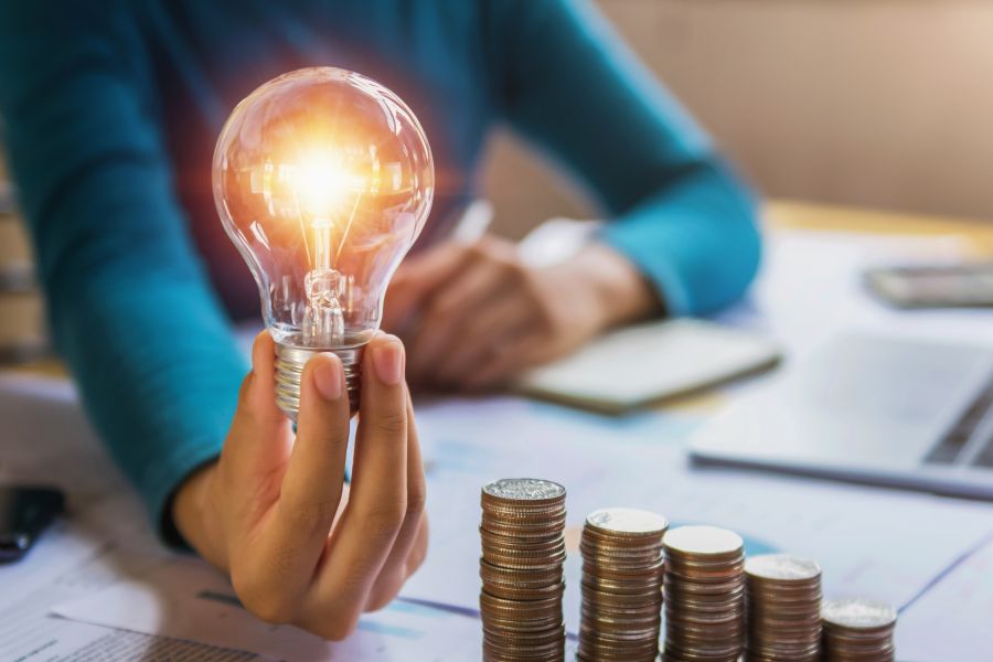Hand holding illuminated light bulb, alongside a pile of coins to indicate investment.
