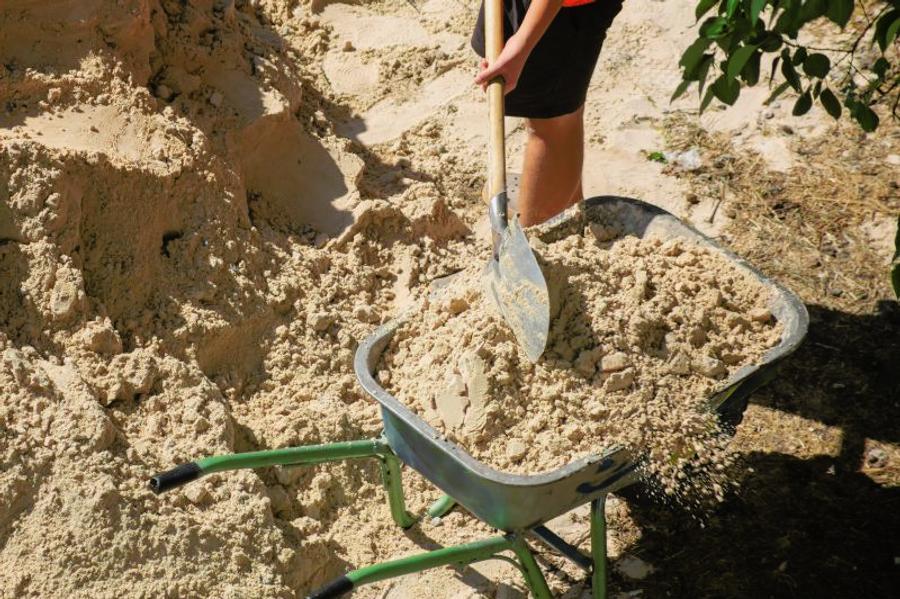 Worker fills wheelbarrow with sand using shovel close-up, sand at construction site.