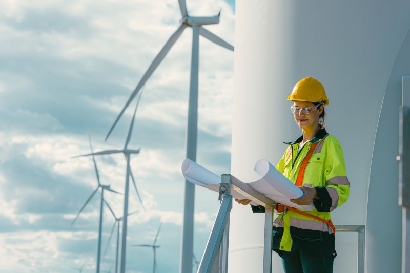 female engineer working outdoor with safety at wind turbines clean energy power station background.