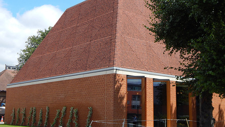 A squat re brick building with a sloped roof and four large windows on the front. A line of tree saplings runs down the side of the building.
