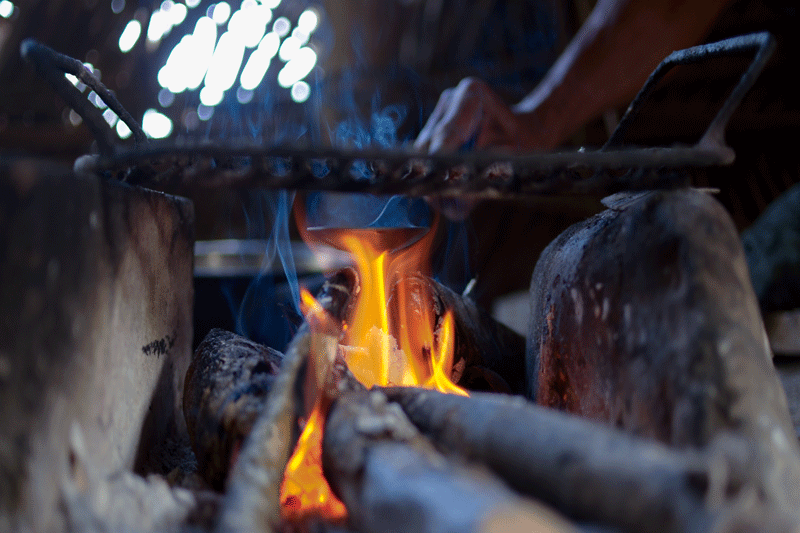 A fire underneath a grill with someone cooking in the background
