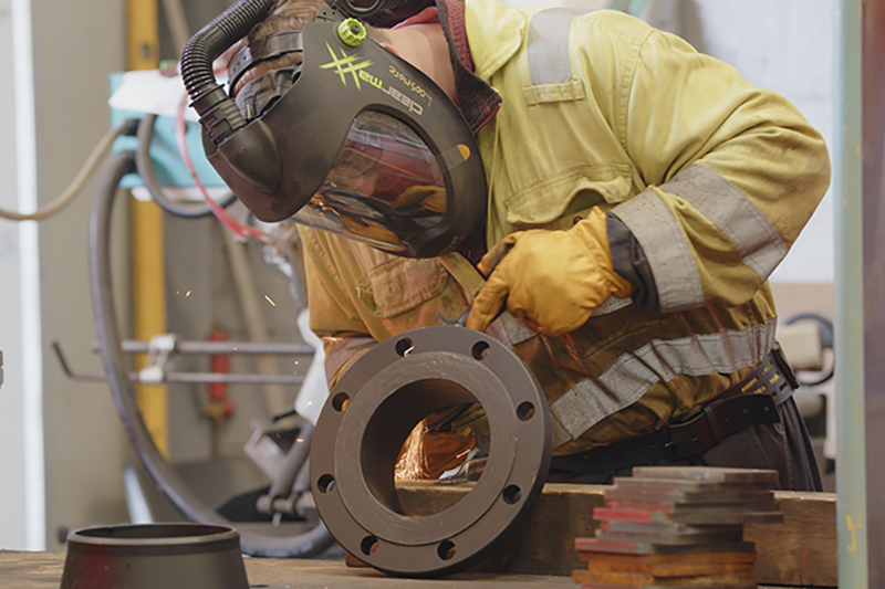 Man in yellow overalls with a mask pointing to round metal object