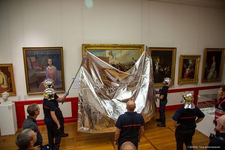 Fire fighters in silver helmets are using long poles to put a silvery blanket over a large painting. Other paintings line the walls