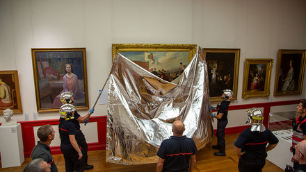 Fire fighters in silver helmets are using long poles to put a silvery blanket over a large painting. Other paintings line the walls