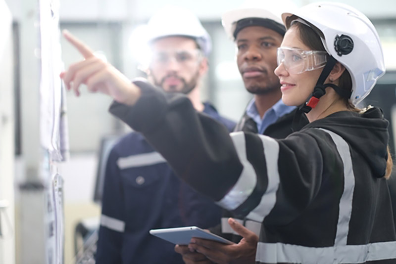 Three young engineers in protective gear have a discussion around a whiteboard