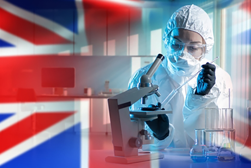 A scientist in full protective gear with goggles using a microscope in front of the Union Jack flag