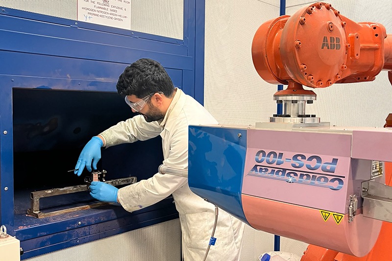 A man in a lab coat, goggles and rubber gloves adjusting an object in a recess in a wall with a very large metal spray machine aimed at it