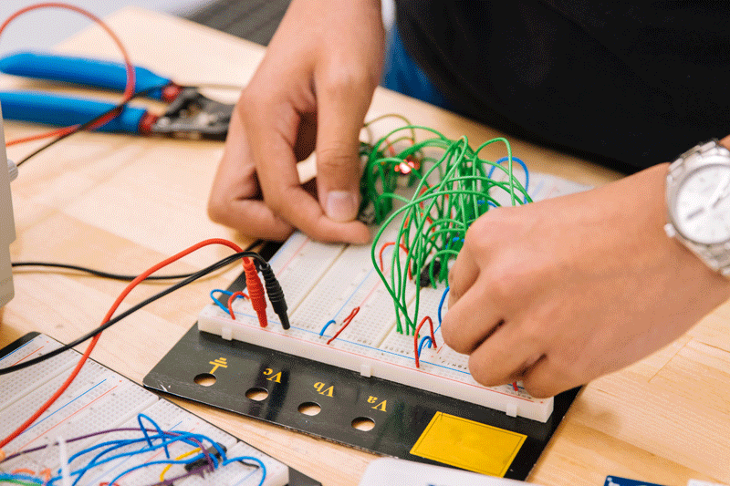 hands at workbench holding green wires on a curcuitboard, it is connected to a supply or output and there are pliers to the right