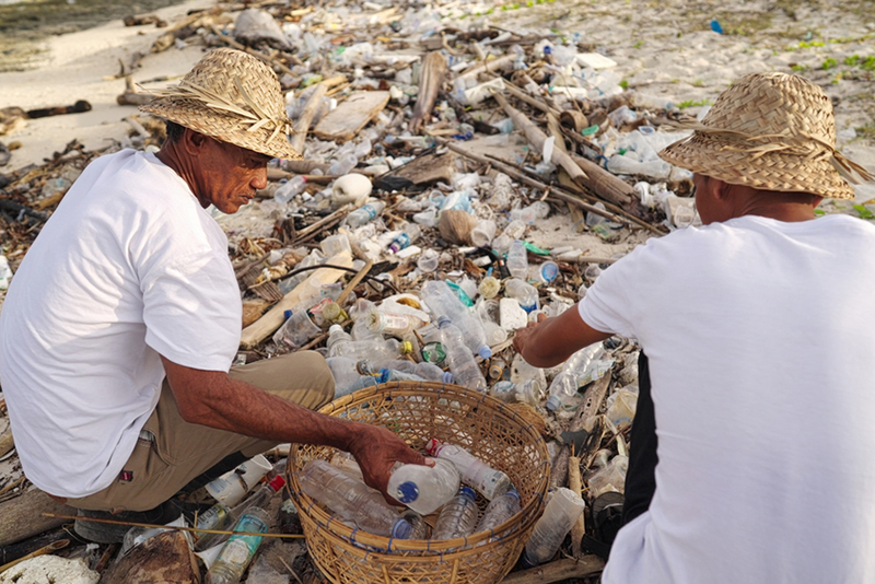 Two plastic waste pickers or collectors collecting bottles on a littered beach in Indonesia for recycling in Indonesia