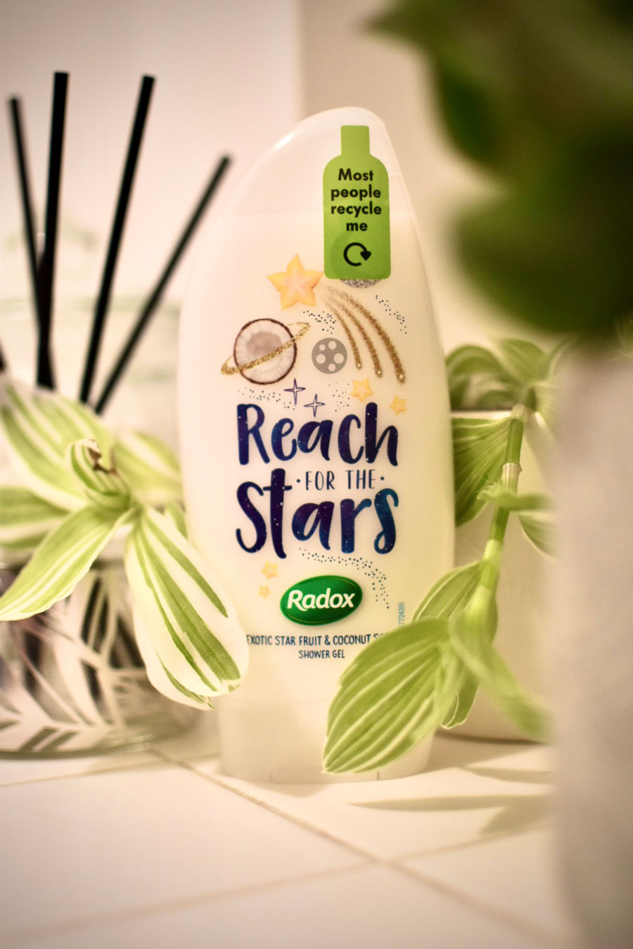 bottle of shampoo with the message "most people recycle me" printed on it, standing on a tiled surface next to a green leafy plant and a diffusor