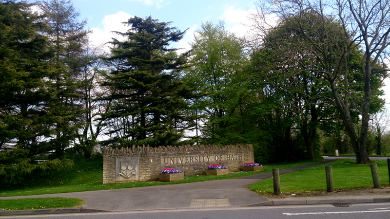 A wall with the words 'University of Bath' and the university coat of arms engraved on it. Planters with flowers in front and a line of trees behind.