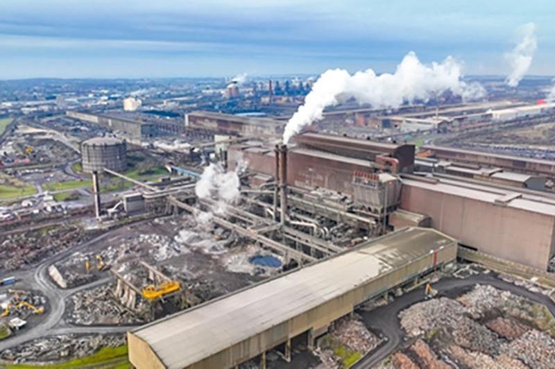 An aerial view of a very large factory with two huge warehouses perpendicular to each other with large chimneys and pipework, with lots of industrial rubbish lying around