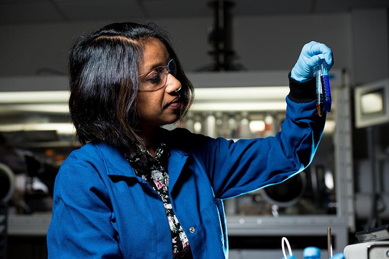 researcher in a lab, wearing goggles, gloves, a blue, open, lab coat and patterned blouse holds up two test tubes and looks at them