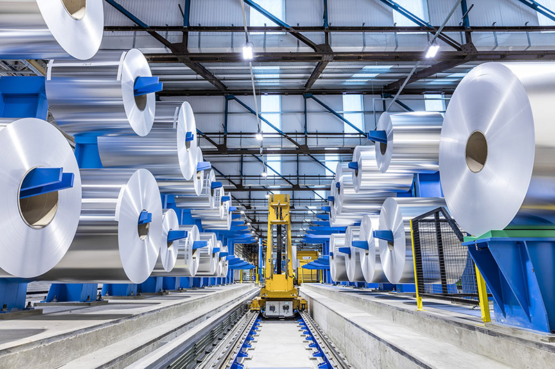 Large metal rolls in a warehouse either side of a small rail track with a mini haulage vehicle in the distance