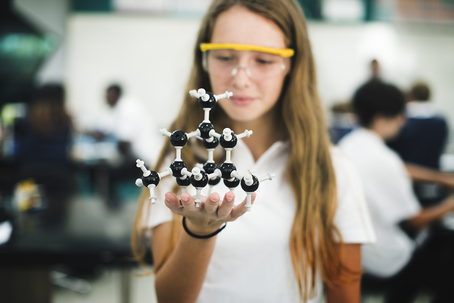 High school student holding a model molecule structure in class, she has goggles, long hair and a white blouse