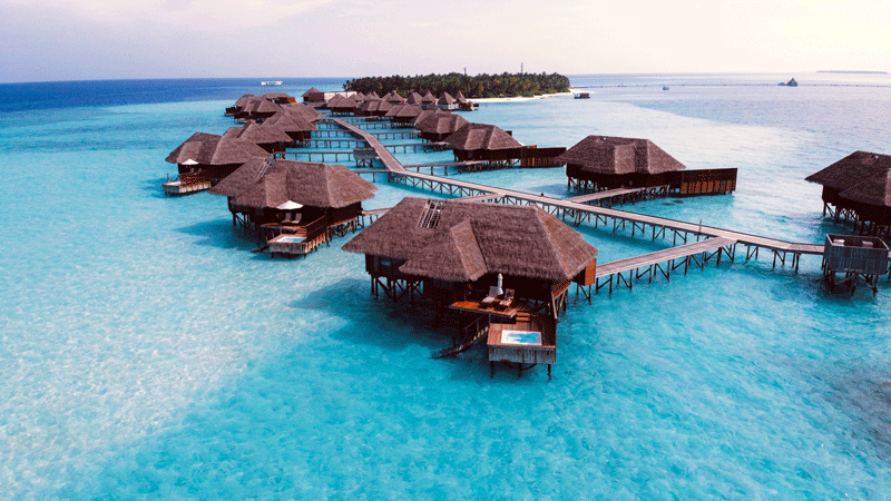 Holiday cabins with boardwalk and mini-pools against a clear light blue sea, a wooded island is in the distance
