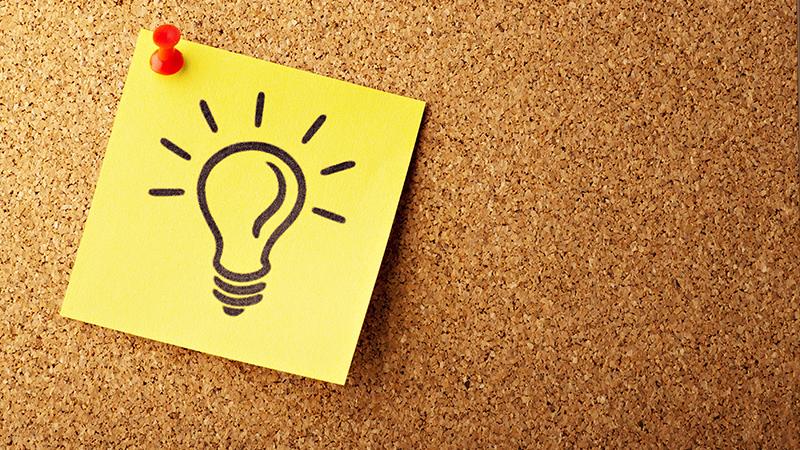 Corkboard with a yellow square of paper stuck on with a red pin, there is a black drawing of a screw in lightbulb with rays of light on the note
