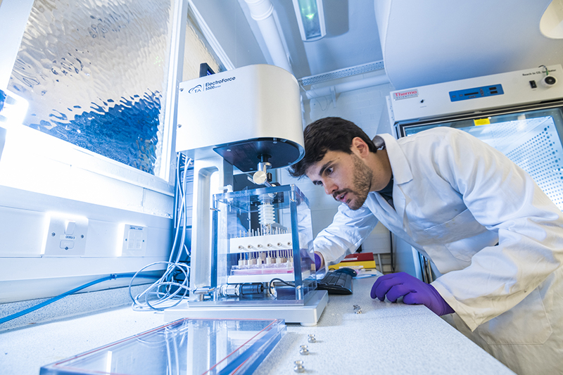 A man in a white coat leans closely in to observe a machine in a lab