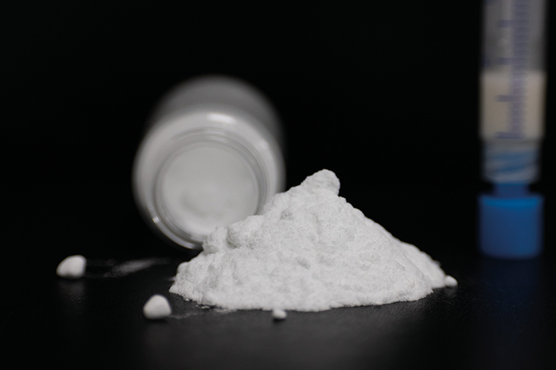 A small pile of white bioglass powder with a vial and syringe behind it, against a black background