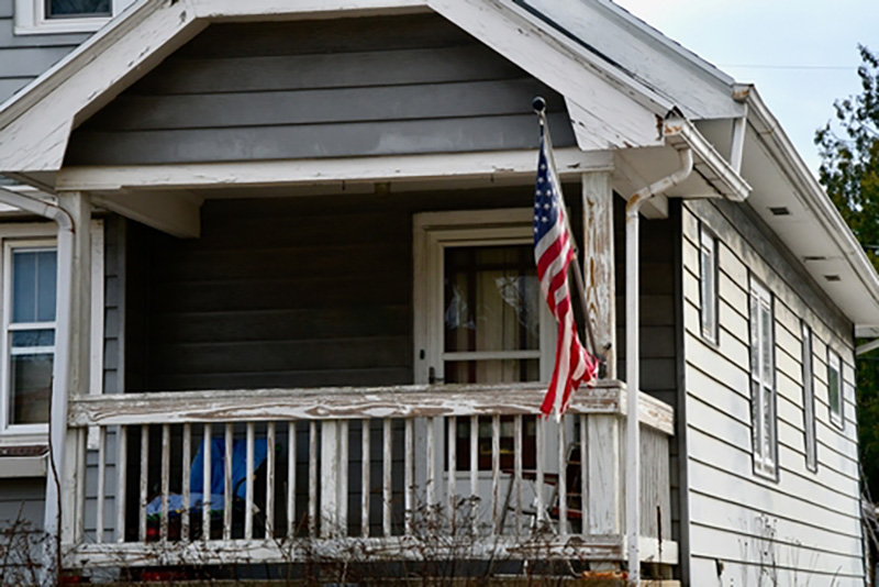 The front of a wooden house with paint flaking off and an American flag attached to the covered porch