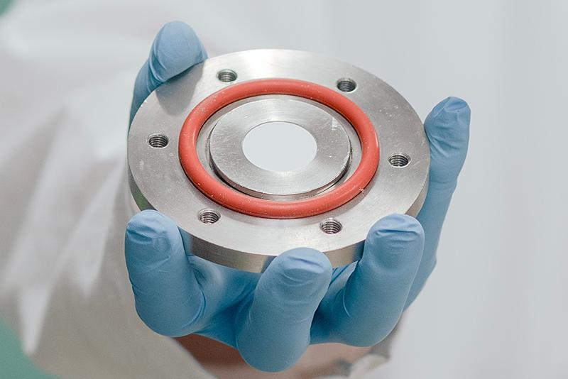 blue gloved hand holds a metal disc with holes around the circumference and an orange ring around the centre, middle has a circular white area