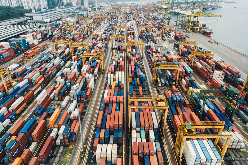 A busy port with many storage containers, orange, white, red and blue on the dockside, one ship is seen on the water by cranes