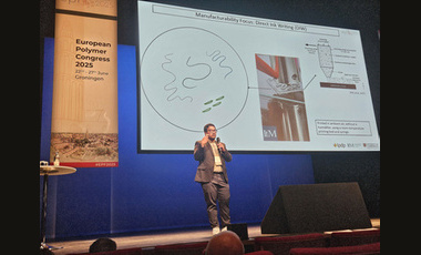 Central large screen showing a presentation, a speaker stands in front of the screen in trousers, glasses and jacket