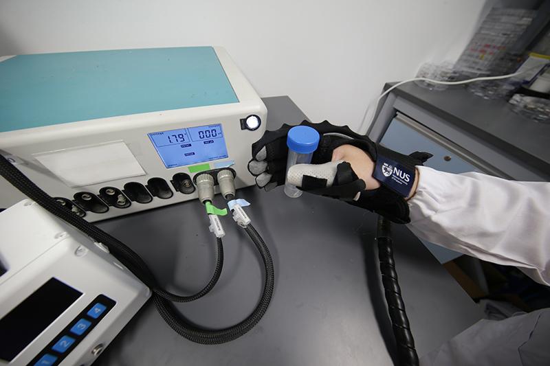 black soft robotic rehabilitation glove on a hand holding a tube with a blue cap sensors on the glove are attached to a reader giving strain figures