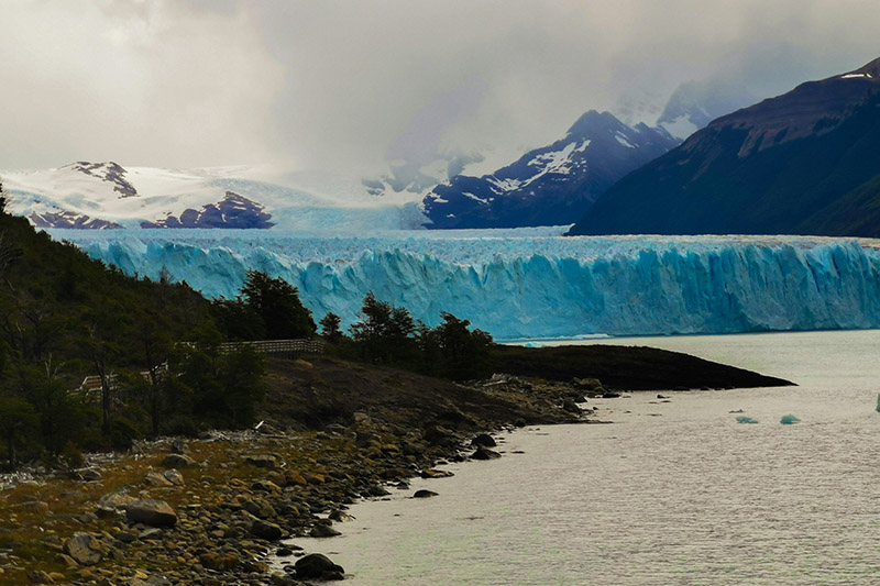 A large wall of ice covering a valley with snowy jagged mountains in the background