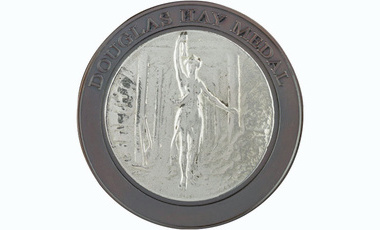 silver boardered circular medal, boarder carries raise text reading Douglas Hay Medal, innet shows goddess with a lamp in a mineshaft