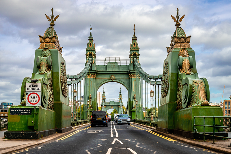 Stock image of one end of the Hammersmith Bridge in London, with a road sign saying 'weak bridge'