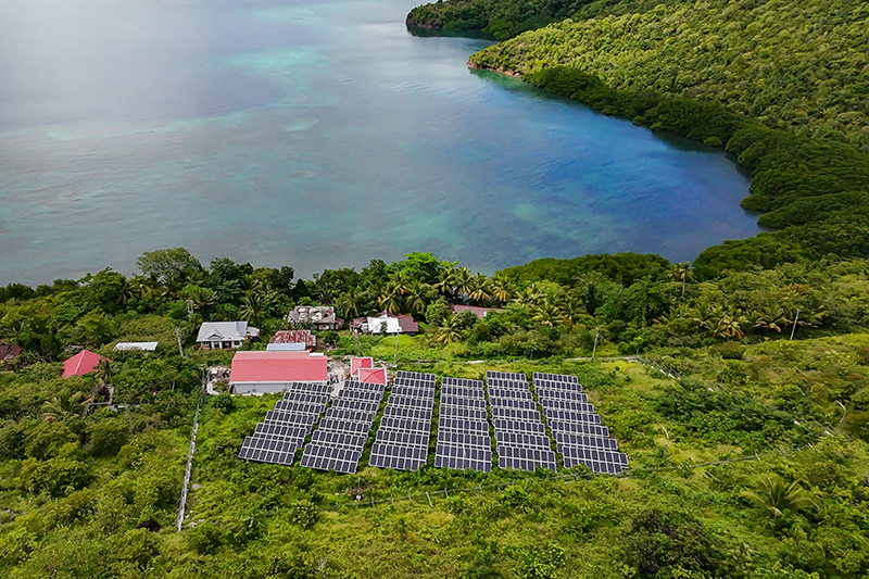 Birds-eye view of solar panels in thick vegetation near two small houses near a large body of water